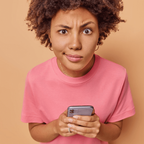 woman concentrated attentively at the camera holds her mobile phone unable to send messages on website form wears a casual pink t-shirt