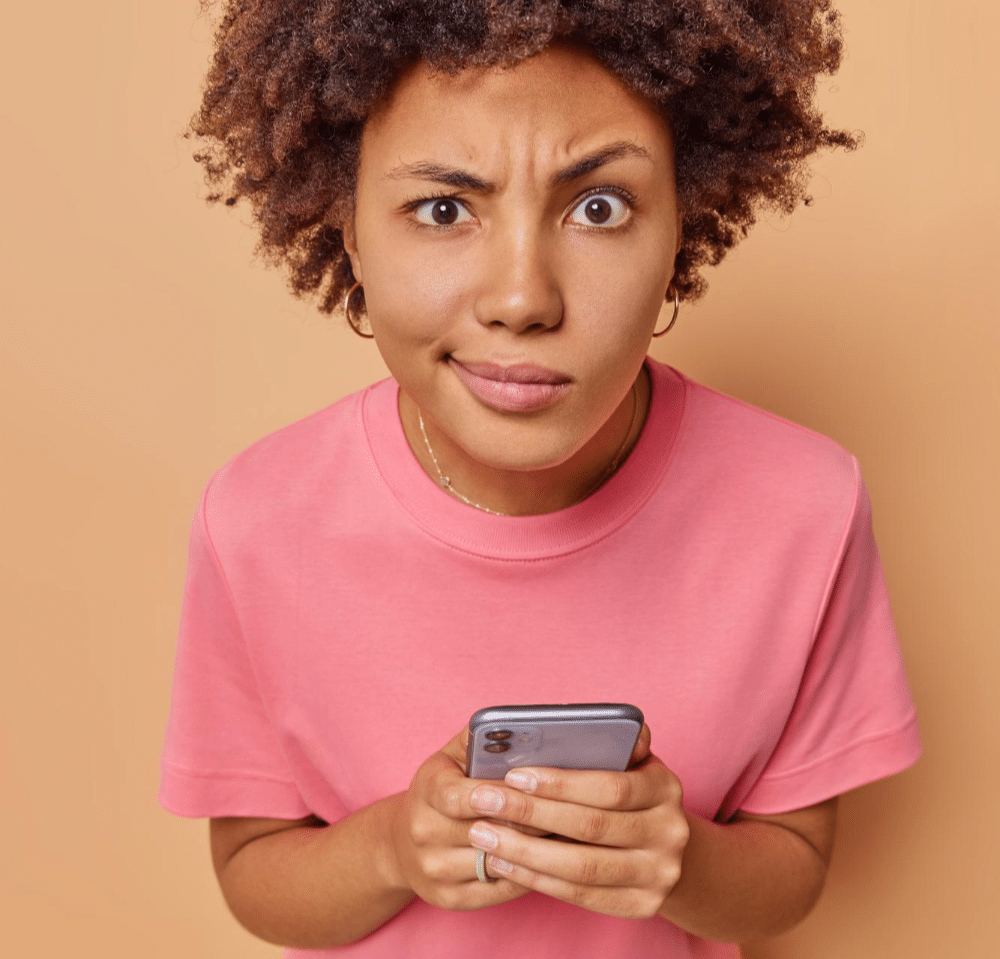 woman concentrated attentively at the camera holds her mobile phone unable to send messages on website form wears a casual pink t-shirt